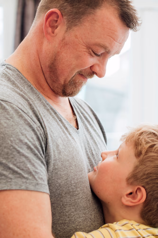 Man in gray shirt smiling down at young boy hugging him while looking up.