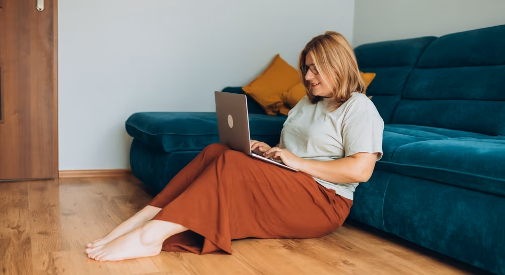 Woman with glasses sitting on floor by teal sofa, using laptop on her lap and smiling.