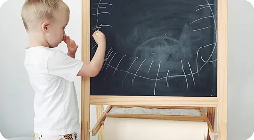 Young boy drawing a smiley face on a blackboard in a bright room.