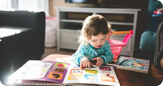 Toddler in blue pajamas reading a colorful picture book on a wooden table in a cozy living room.