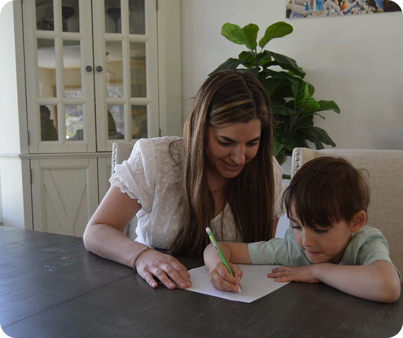 Woman helping young boy write on paper at a dark wooden table inside a home.