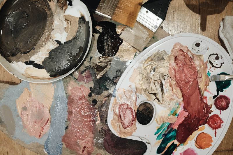 Artist’s palette and bowls with various mixed oil paint colors including beige, brown, pink, red tones, and brushes on a wooden surface.