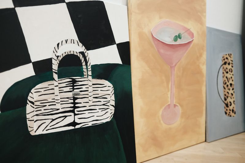 Three artistic paintings showing a zebra-striped handbag on a green surface in front of a black and white checkered background, a pink cocktail glass with two green olives on a beige background, and a leopard print handbag handle on a gray background.