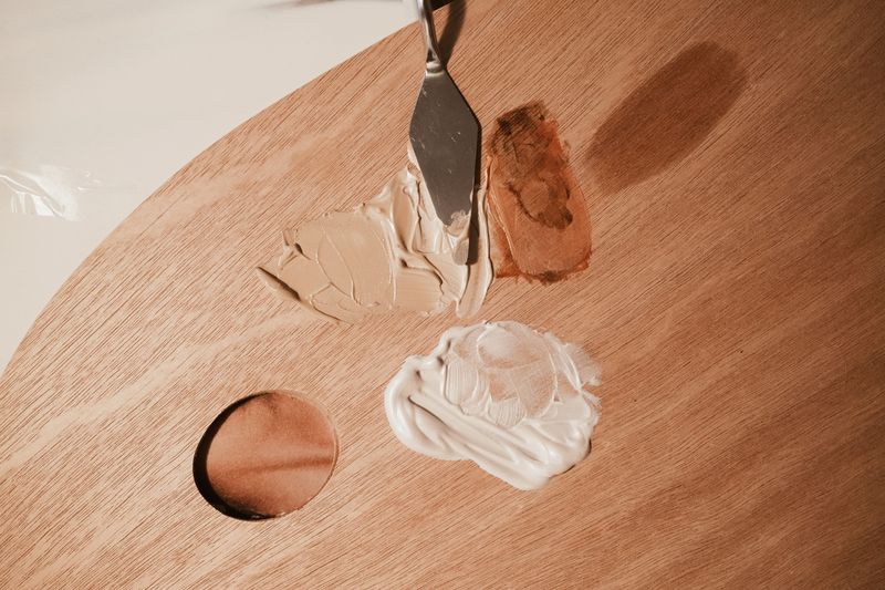 Palette with different shades of beige and brown cream makeup, with a metal spatula spreading one shade.