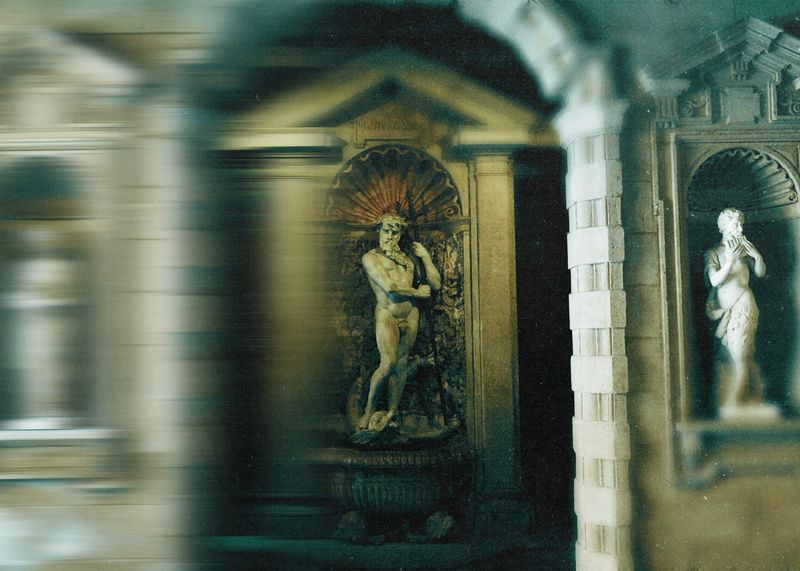 Sculpture of a nude male figure with a beard standing in an alcove next to a sculpture of a partially draped female figure, both illuminated against a dark background in a classical architectural setting.
