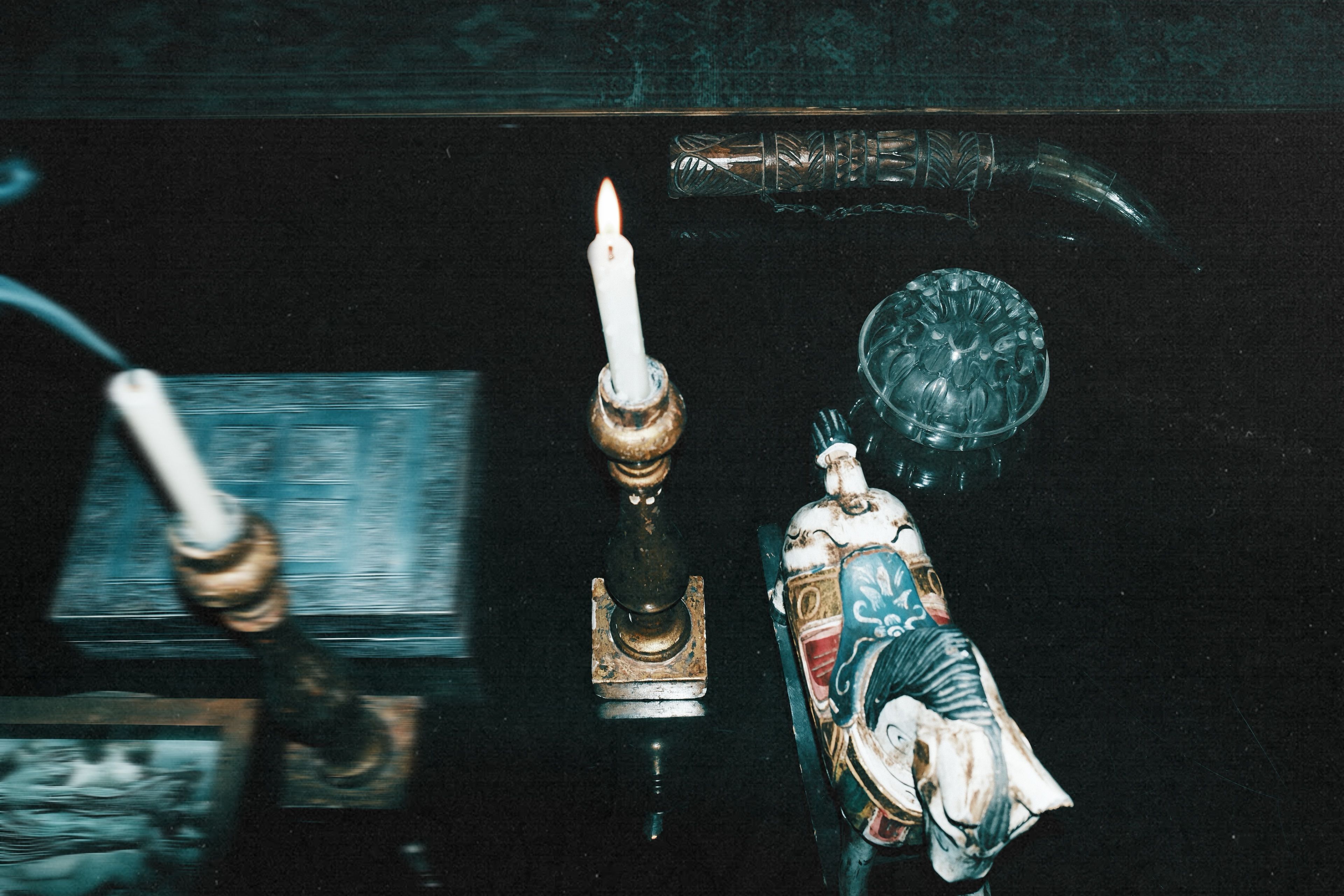 Dark reflective surface with a lit candle in a brass holder, a decorated horse figurine, a glass bowl, and an ornamental curved object.
