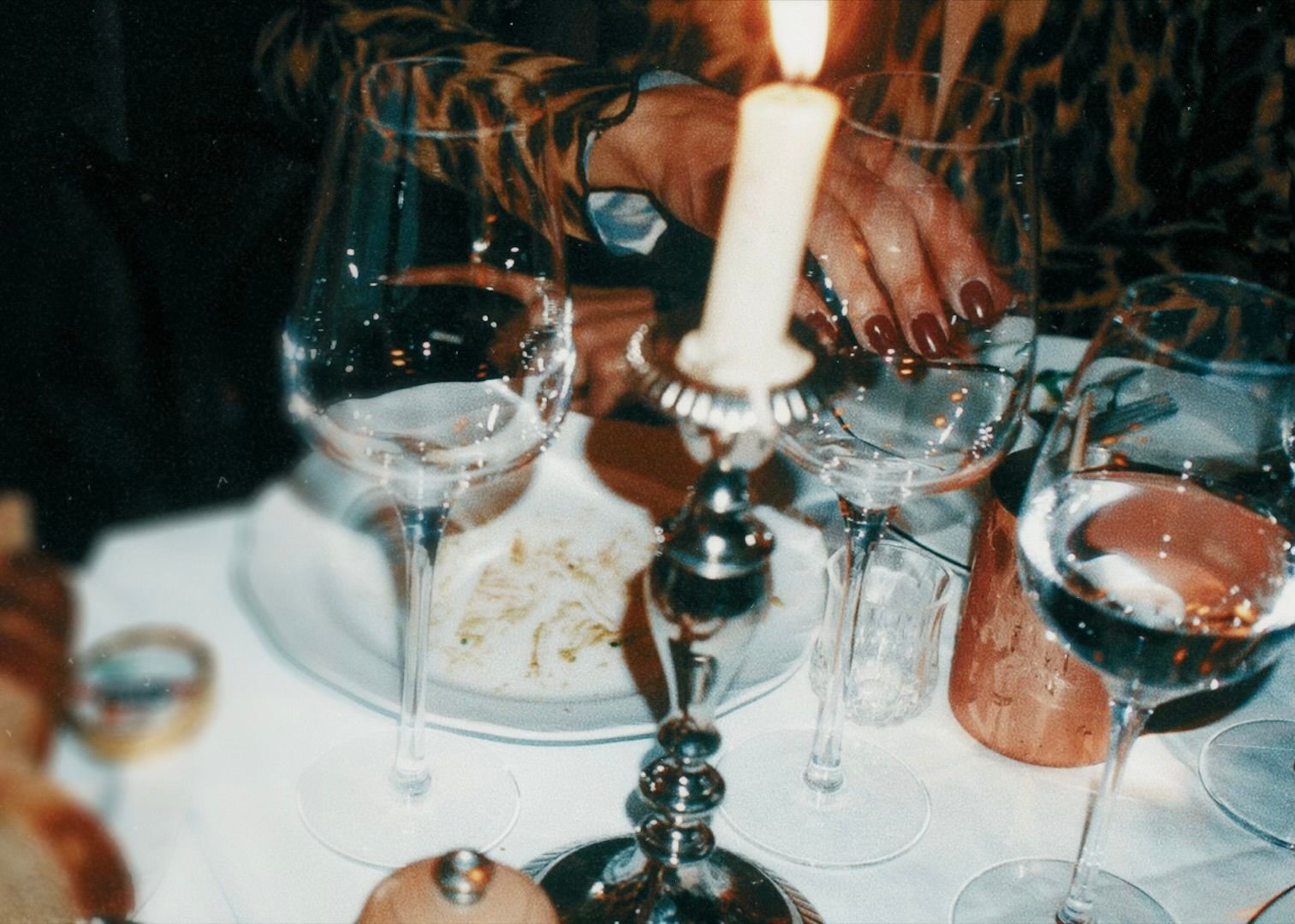 A hand with red nail polish reaching over a table set with wine glasses, a lit candle in a silver candlestick, and a white plate with some food remnants.