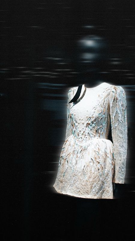 White long-sleeve dress with intricate textured pattern displayed on a black mannequin against a dark background.