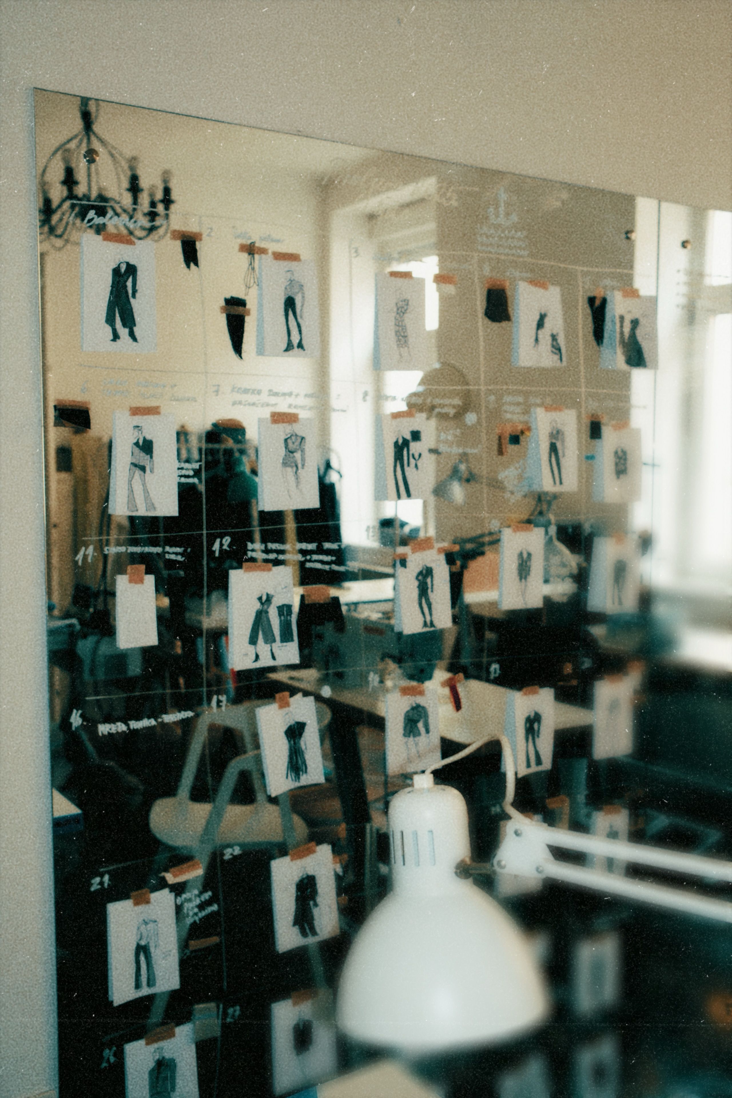 Reflection of a workspace with a blackboard covered in fashion sketches of clothing designs and a white desk lamp in the foreground.