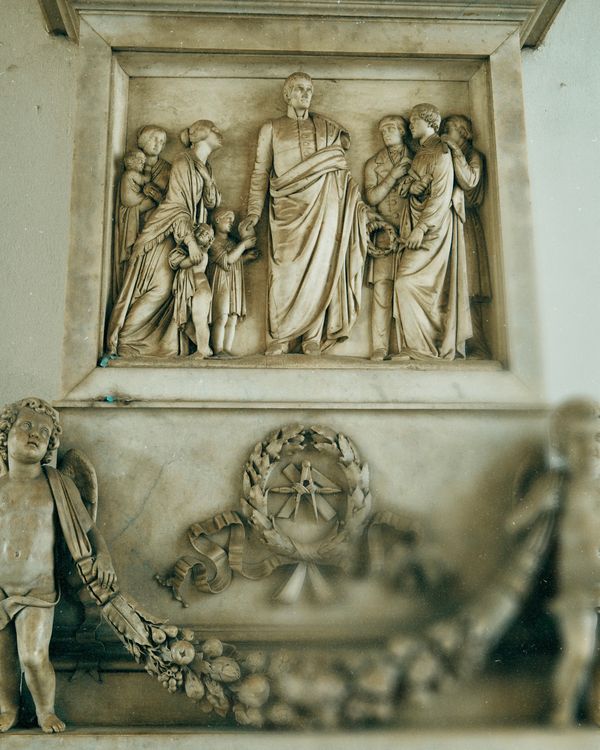Marble bas-relief sculpture depicting a central figure surrounded by men, women, and children in classical robes, with two cherubs holding garlands below.