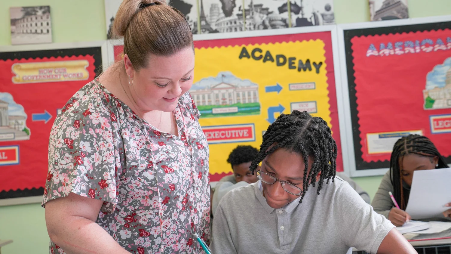 Teacher assisting a student writing with a pencil in a classroom with educational posters