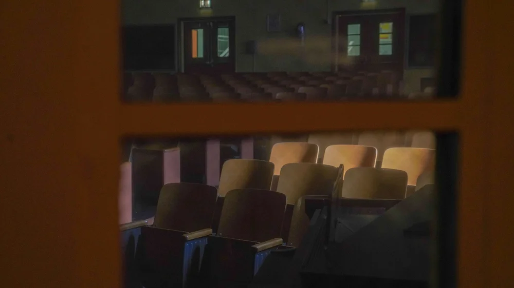 Empty auditorium with rows of brown chairs viewed through a wooden window.