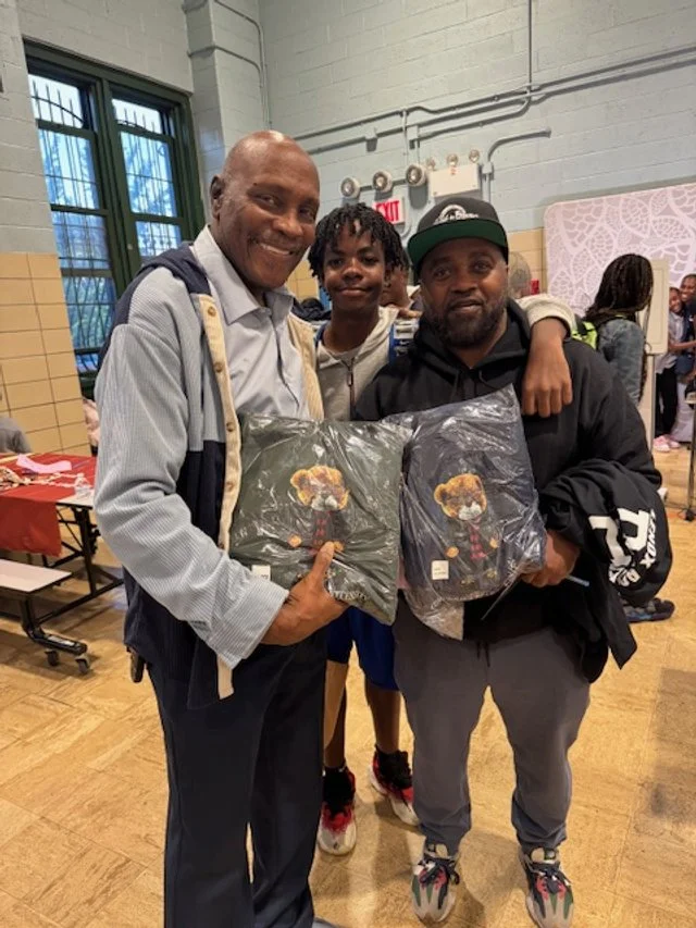 Three smiling men standing indoors, two holding packaged clothing with teddy bear designs.