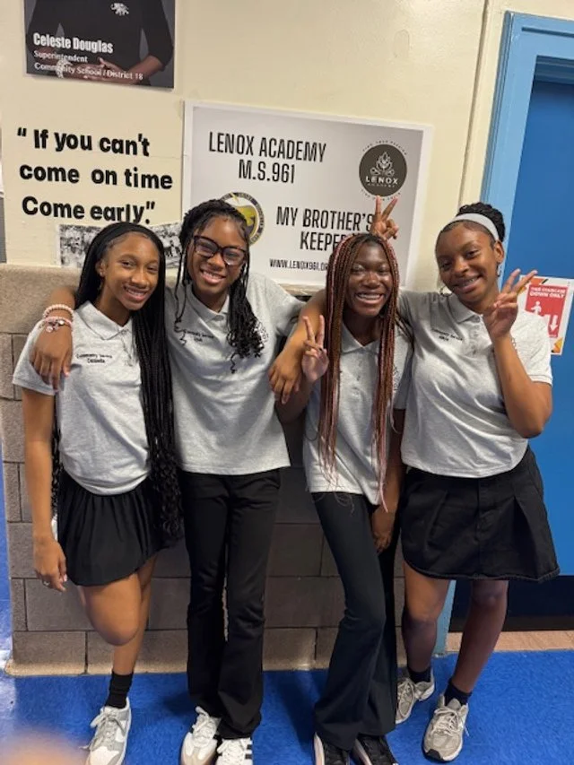 Four smiling teenage girls in school uniforms posing together indoors.
