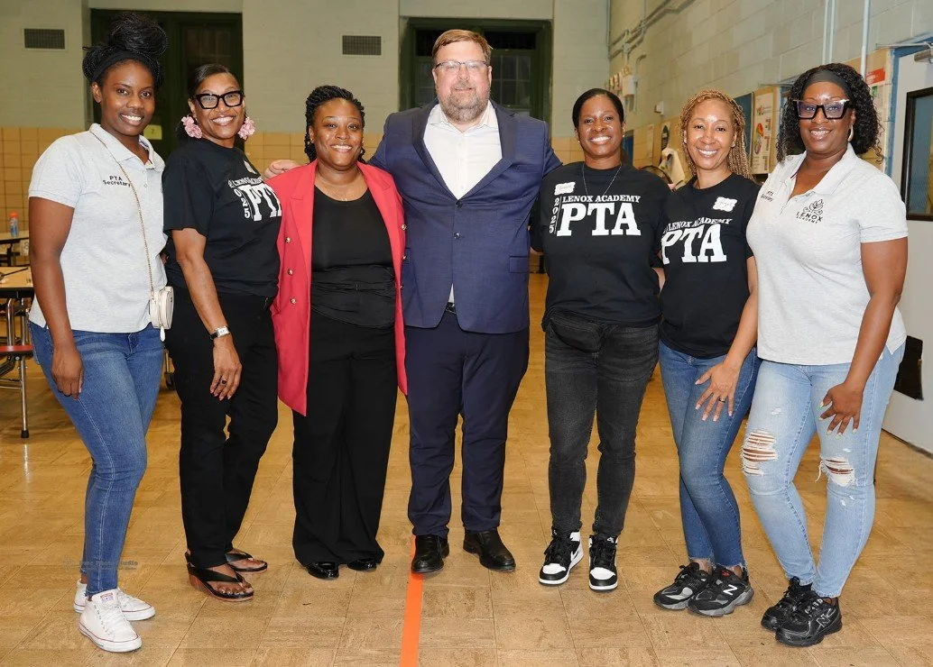 Seven adults standing side by side in a school gymnasium, smiling at the camera
