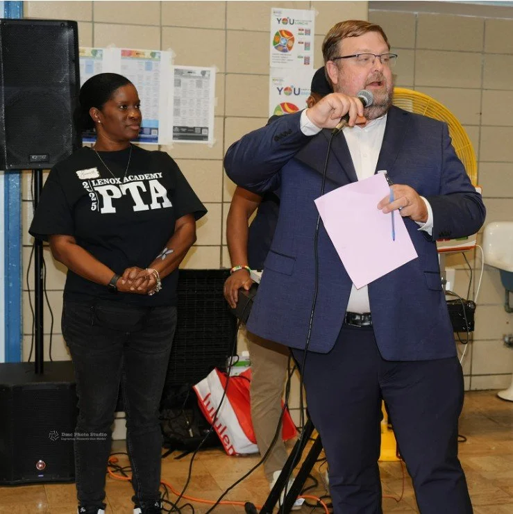 Man in a blue suit speaking into a microphone while holding papers