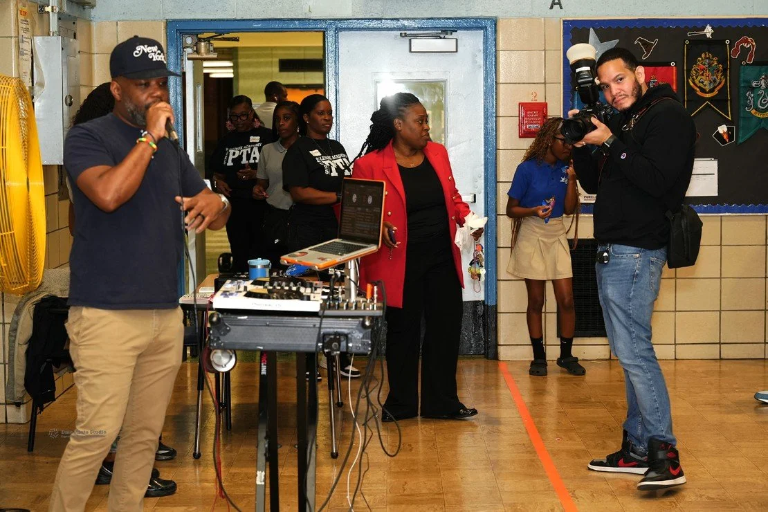 Man in a blue shirt and beige pants speaking into a microphone near DJ equipment