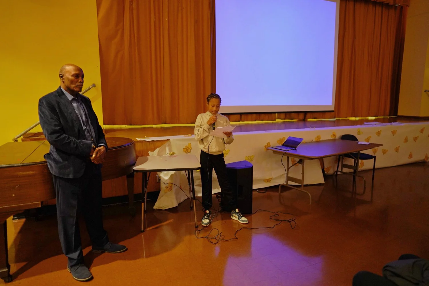 A child stands on a stage holding a microphone and reading from a paper