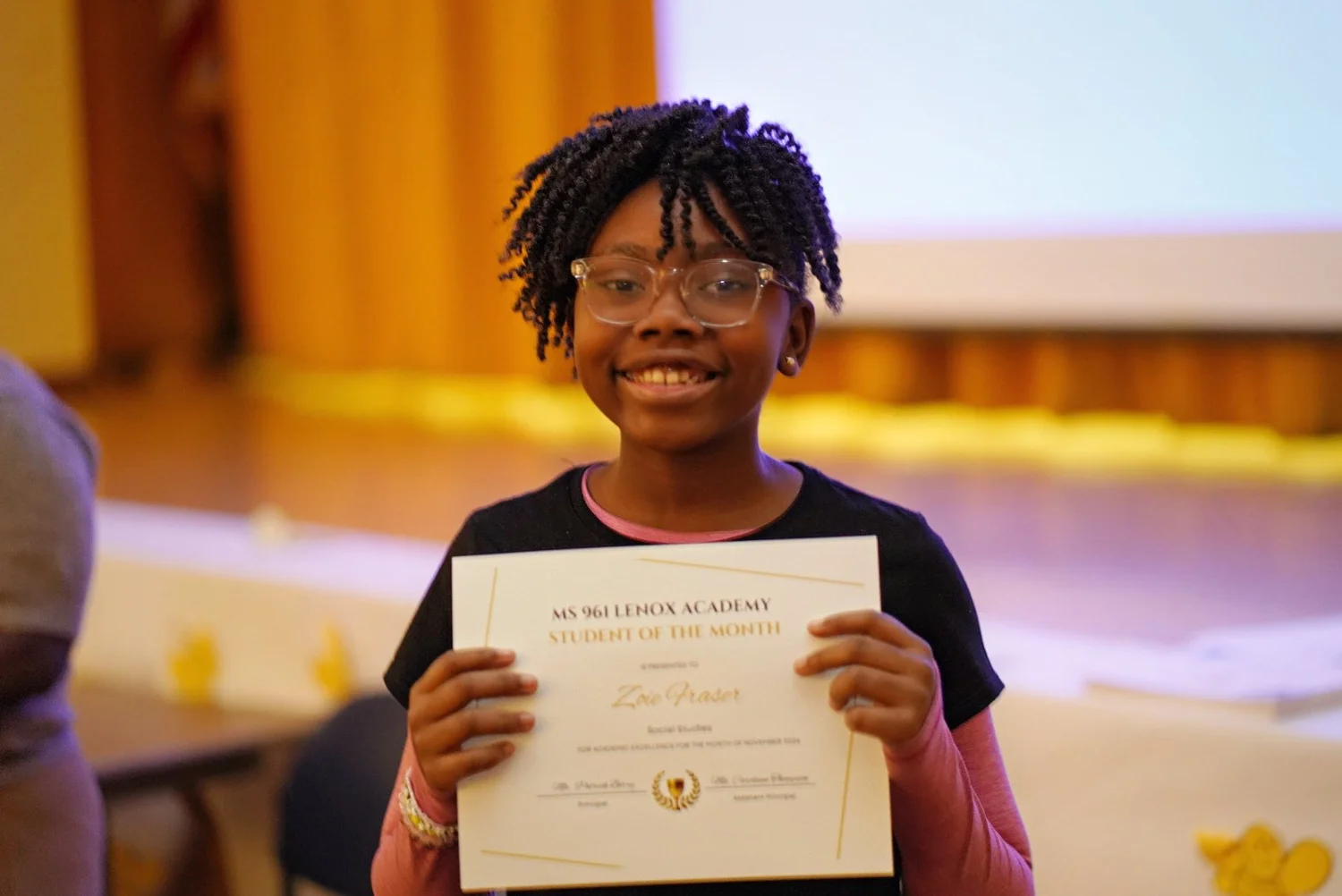 Smiling girl wearing glasses holding a 'Student of the Month' certificate in a school setting.