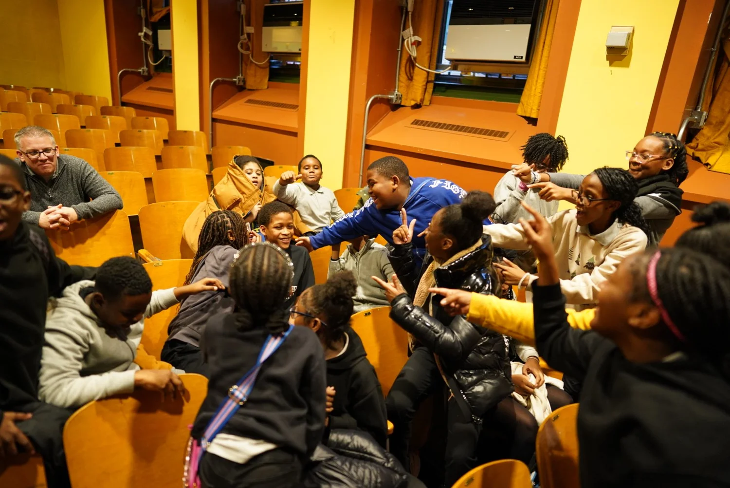 A group of children and one adult sitting in yellow auditorium seats