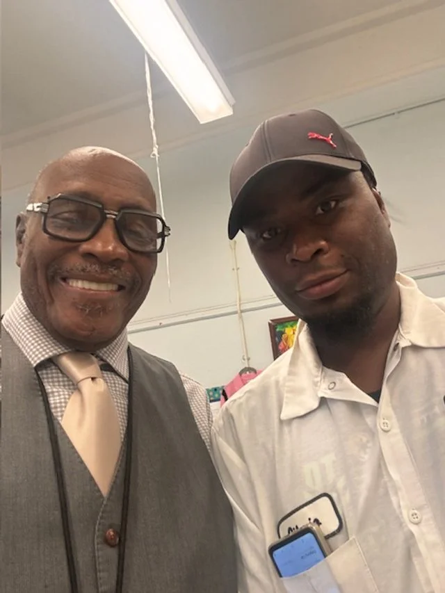 Two men standing indoors, one wearing glasses, a checkered shirt