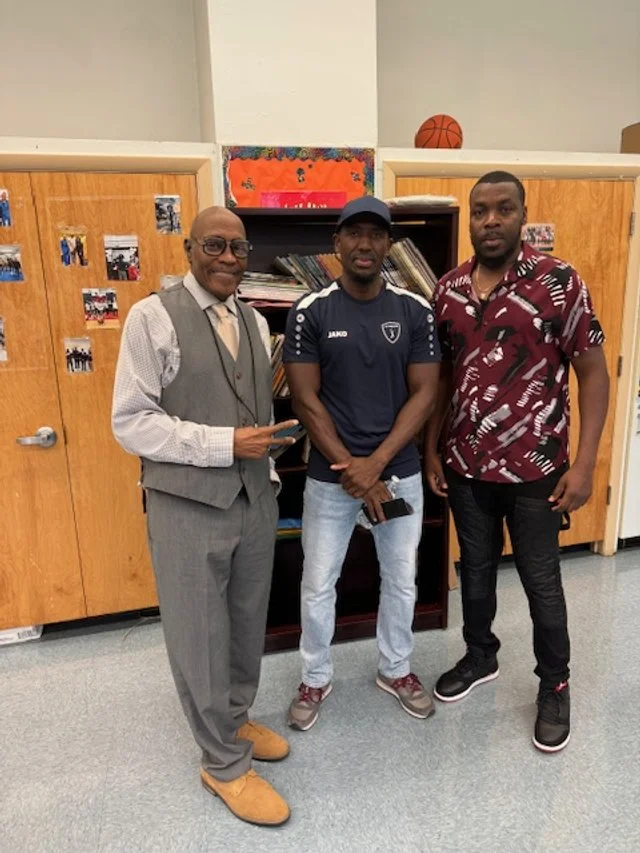 Three men standing indoors in front of wooden cabinets and a bookshelf