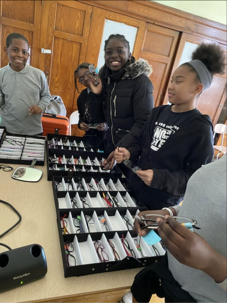 A group of children selecting eyeglasses from organized trays on a table indoors.