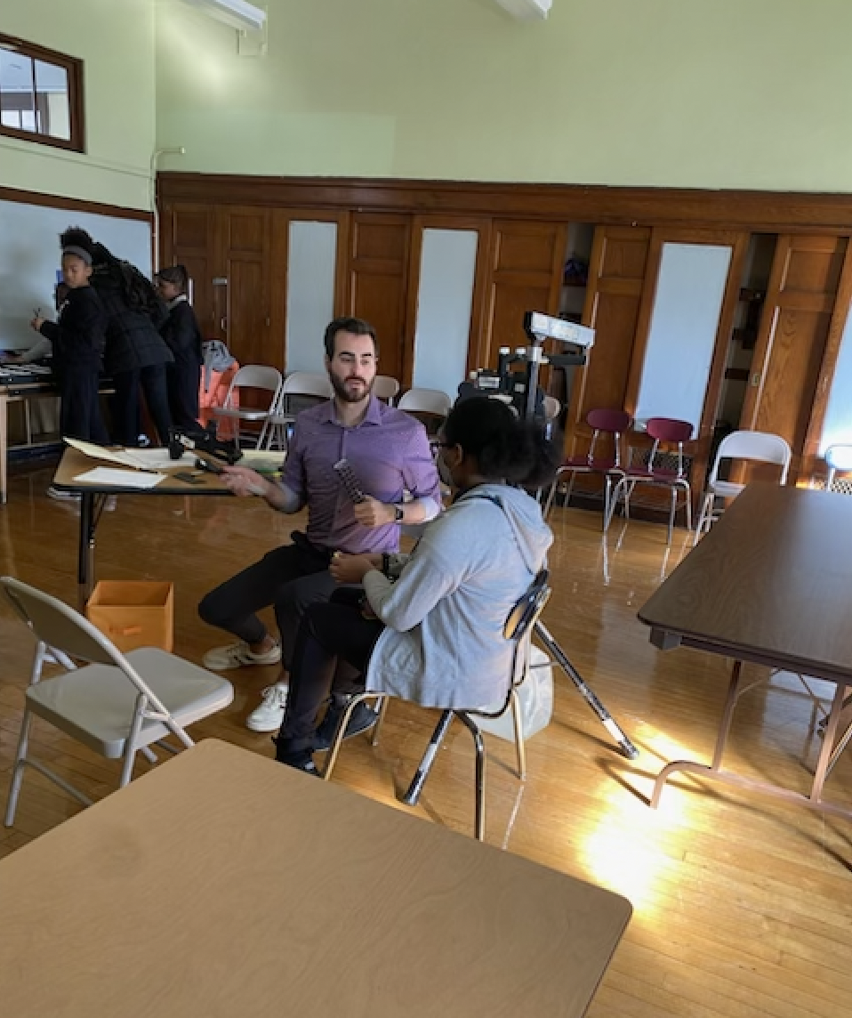 Man in a purple shirt kneeling and talking to a seated woman in a gray hoodie