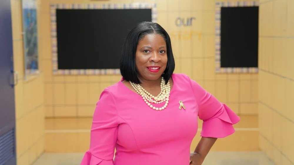 Woman in a bright pink dress with pearl necklace and a pink ribbon pin, standing confidently in a hallway.