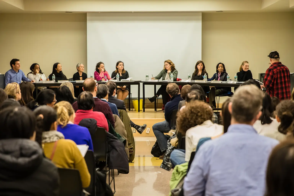 Panel of ten diverse people seated at a long table facing an audience in a conference room.