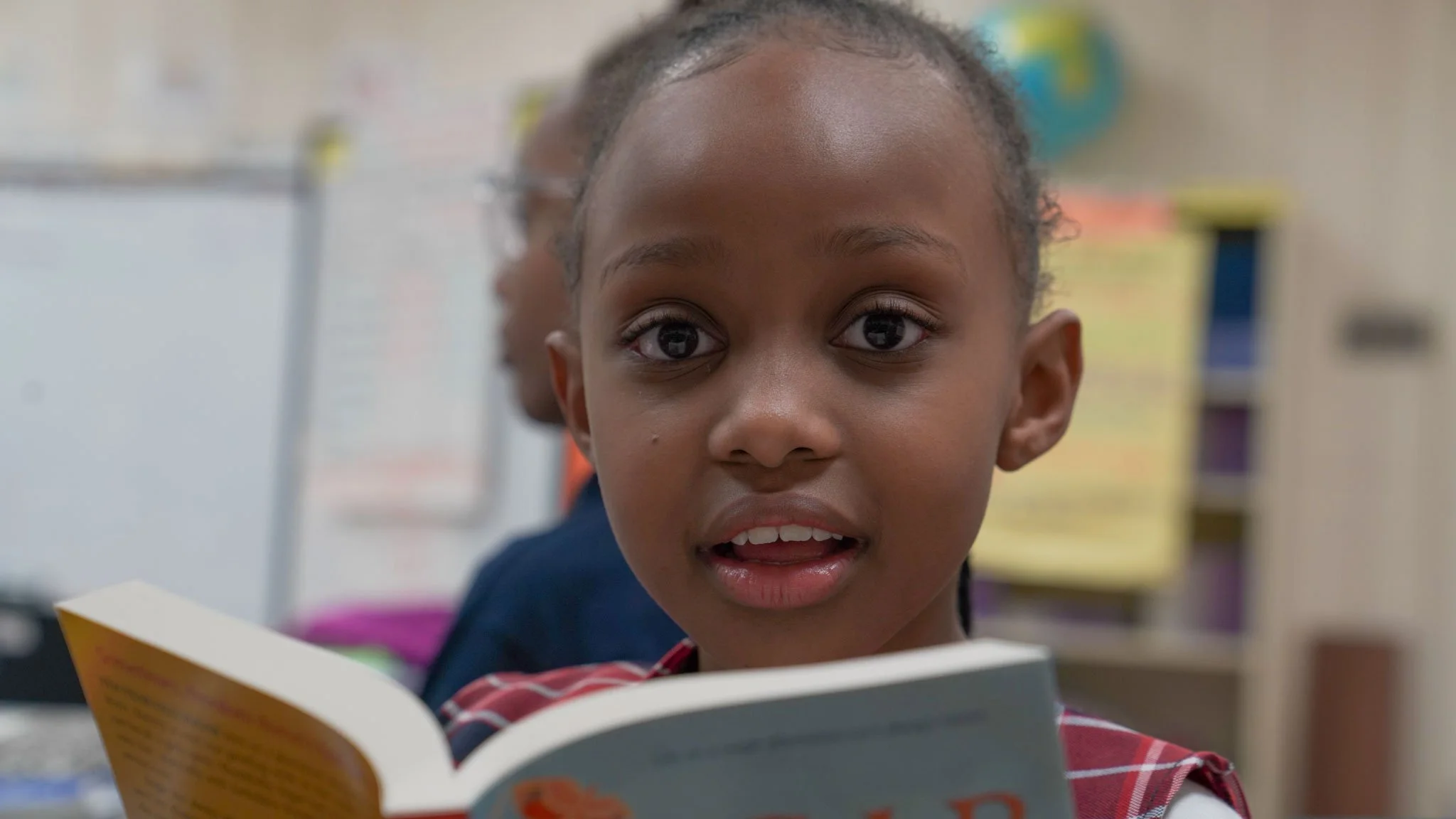 Young girl reading a book aloud in a classroom setting.