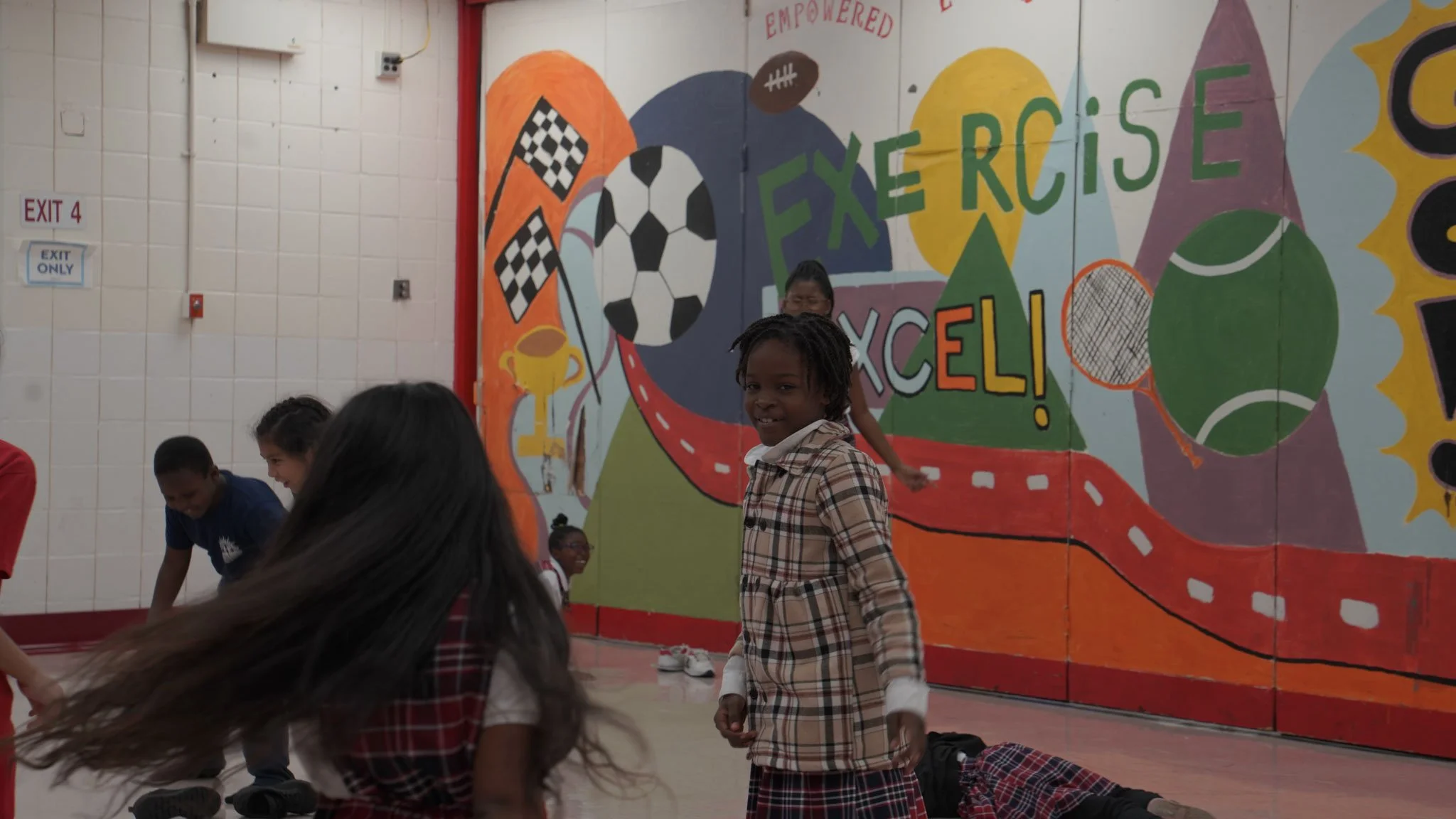 Children playing and smiling in a gym with a colorful mural promoting exercise and sports on the wall.
