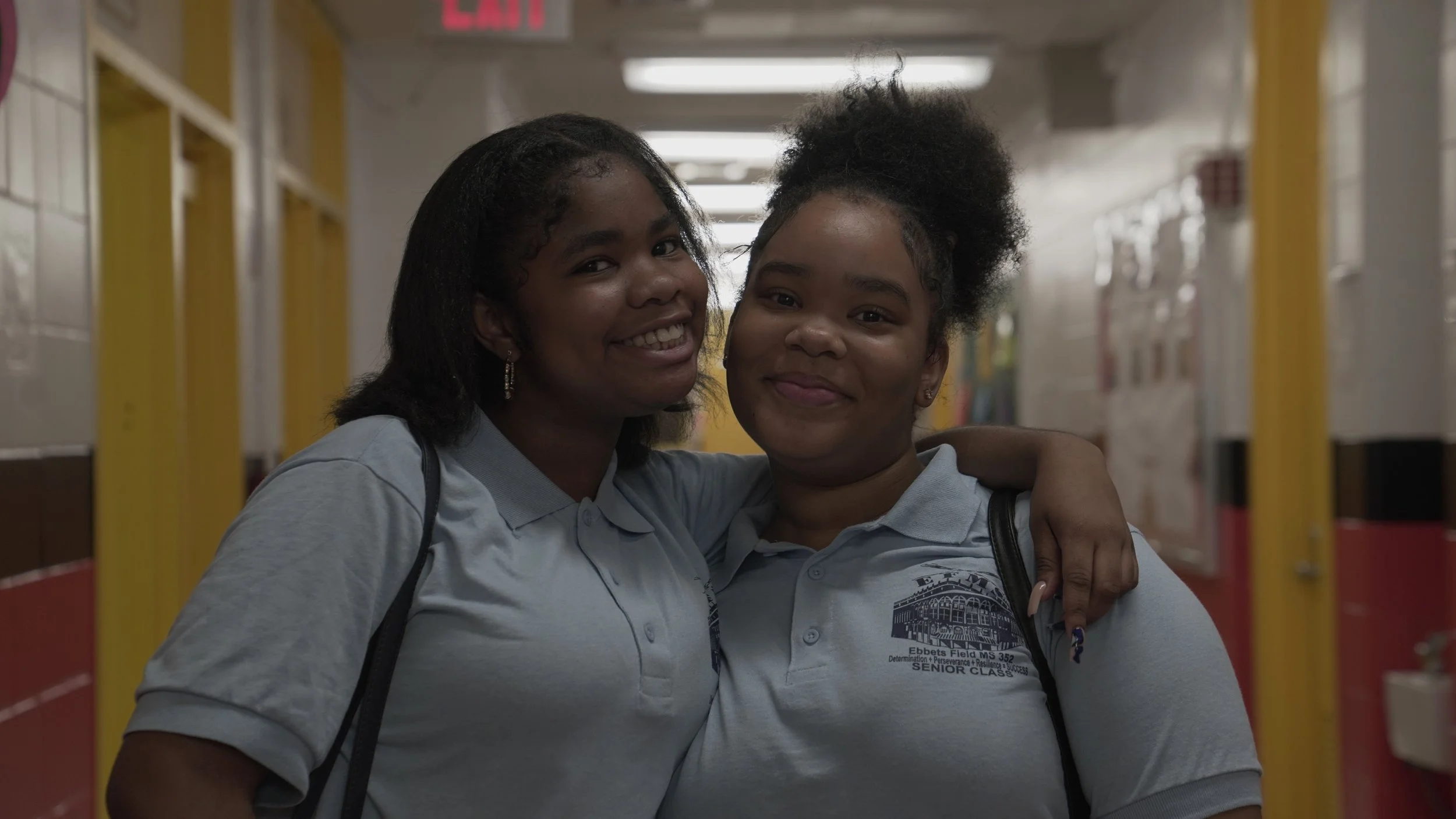 Two young women smiling and hugging in a school hallway wearing light blue polo shirts.