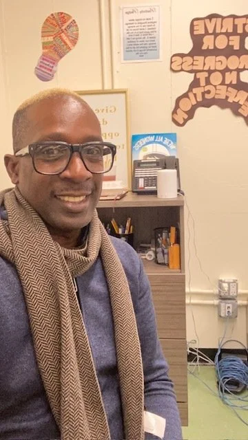 Smiling man wearing glasses and a scarf sitting indoors with office supplies and motivational posters on the wall behind him.