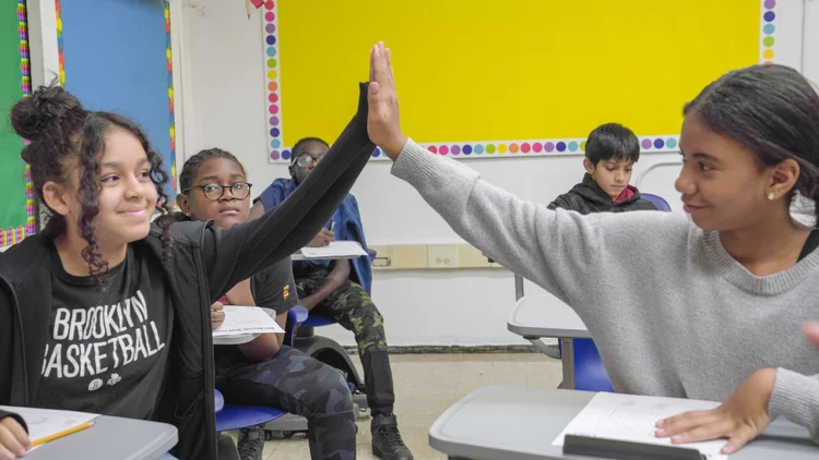 Two students sitting at classroom desks giving each other a high five, with other students in the background.