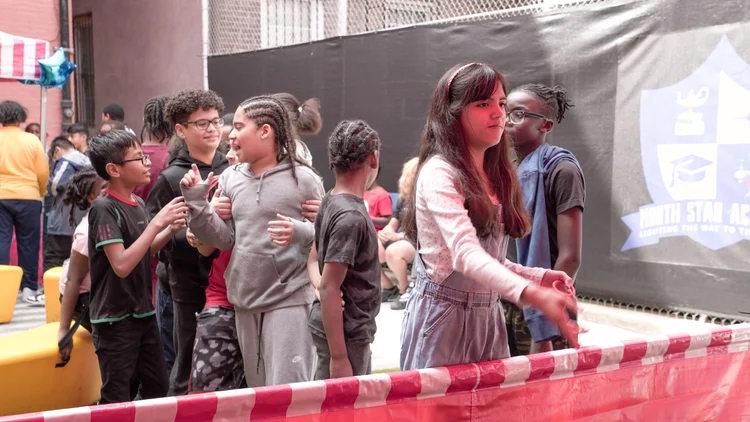 A group of children standing and talking near a red and white striped barrier in an outdoor area with a North Star Academy banner in the background.