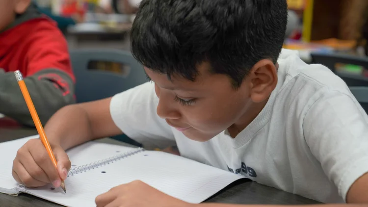 Young boy focused on writing in a spiral notebook with a pencil at a classroom desk.