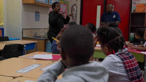 Teacher speaking to young students seated at desks in a classroom.