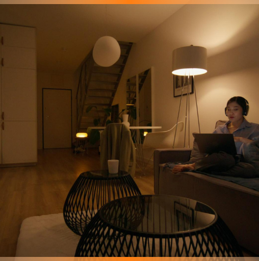 Woman wearing headphones sitting on a couch using a laptop in a dimly lit modern living room with stylish round tables and a floor lamp.