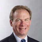 Smiling middle-aged man with short light brown hair wearing a dark suit, white shirt, and blue tie against a light gray background.