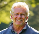 Smiling middle-aged man with curly white hair and sunlit background of green foliage.