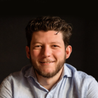 Smiling man with curly hair and a beard wearing a light blue button-up shirt against a black background.