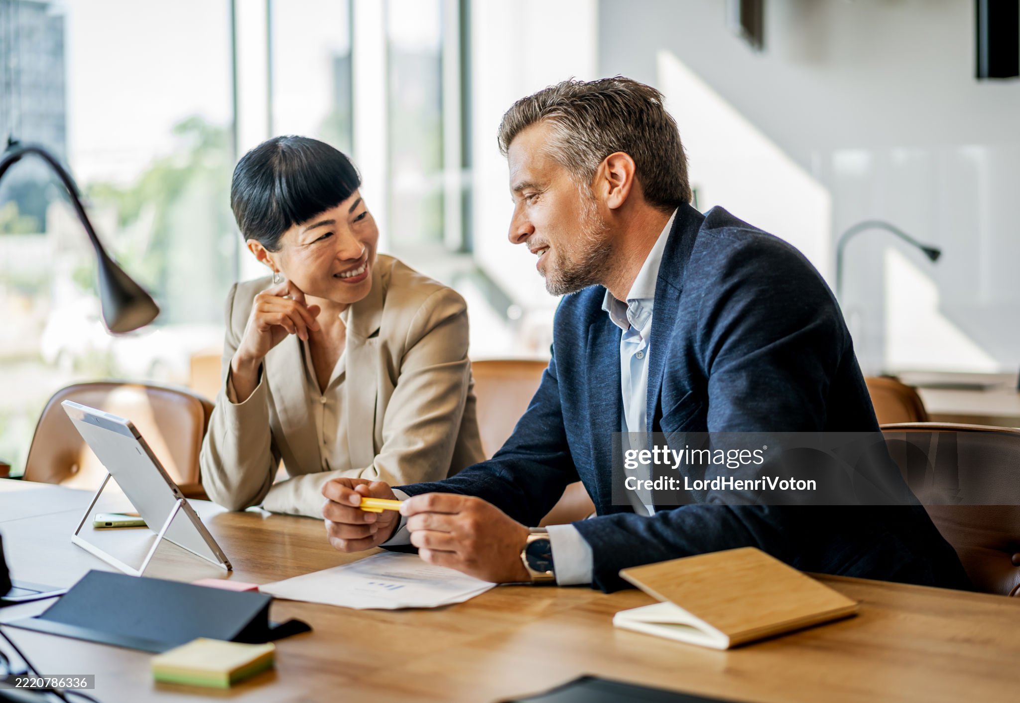 Two business professionals, a woman and a man, sitting at a table engaged in a discussion with documents and a tablet in a well-lit office.
