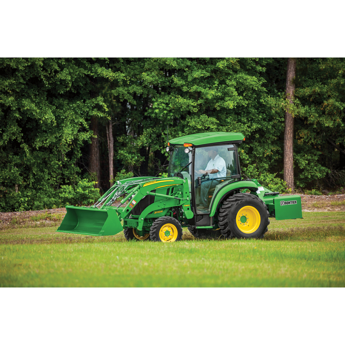 Green tractor with yellow wheels driving across a grassy field, equipped with a front loader bucket and enclosed cab with trees forming a dense backdrop