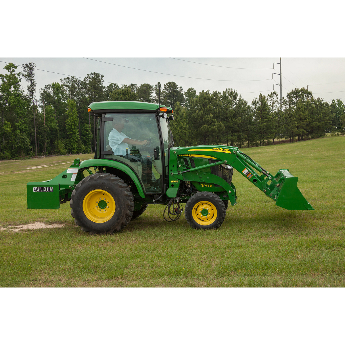 Side view of green tractor with front loader attachment on grass, with an operator inside the cab and trees lining the background under an overcast sky