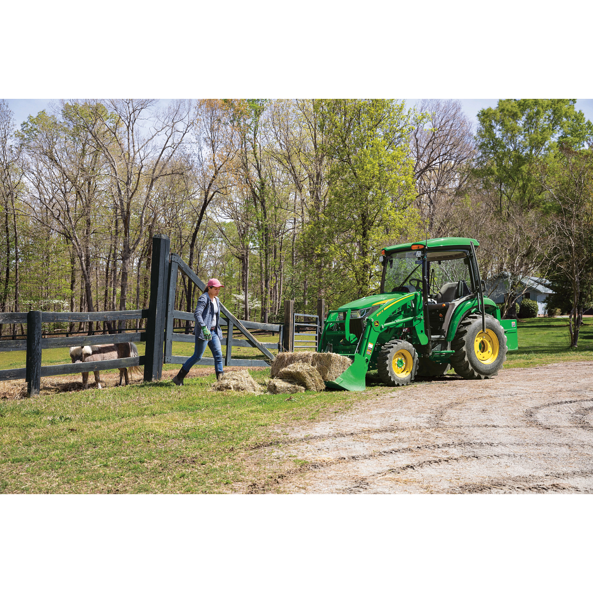 Green 320R tractor parked alongside a horse pen, with driver walking toward it to pick up hay bales in its front loader bucket
