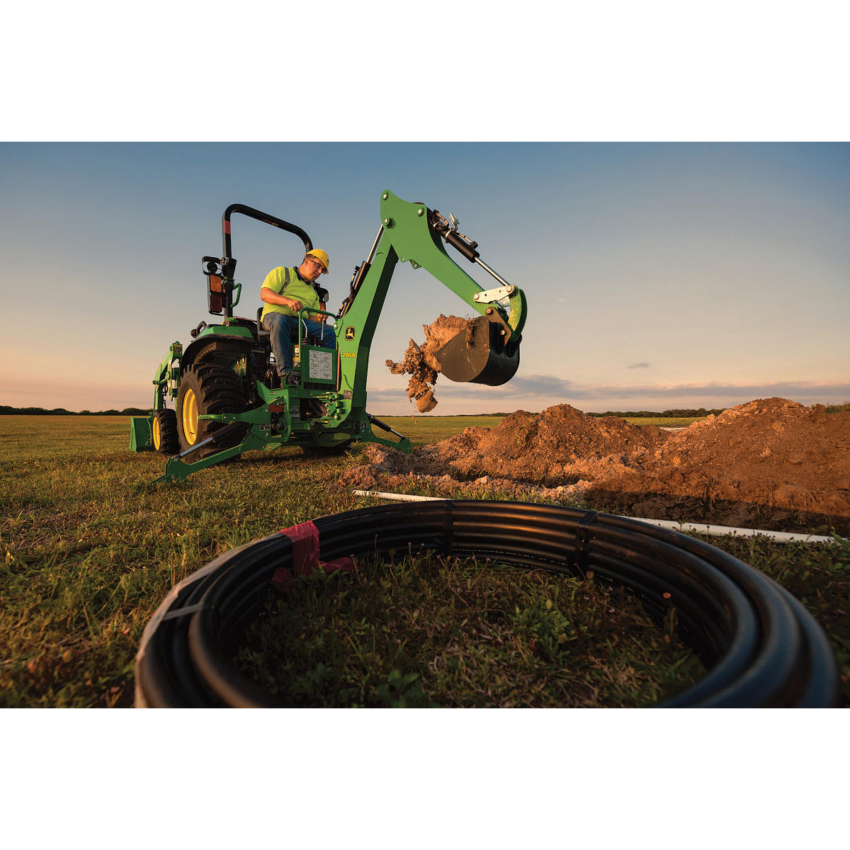 Green compact tractor with a front loader scoops dirt near a circular stack of black pipes in a grassy field at sunset, with a soil mound nearby and warm evening light under a clear sky