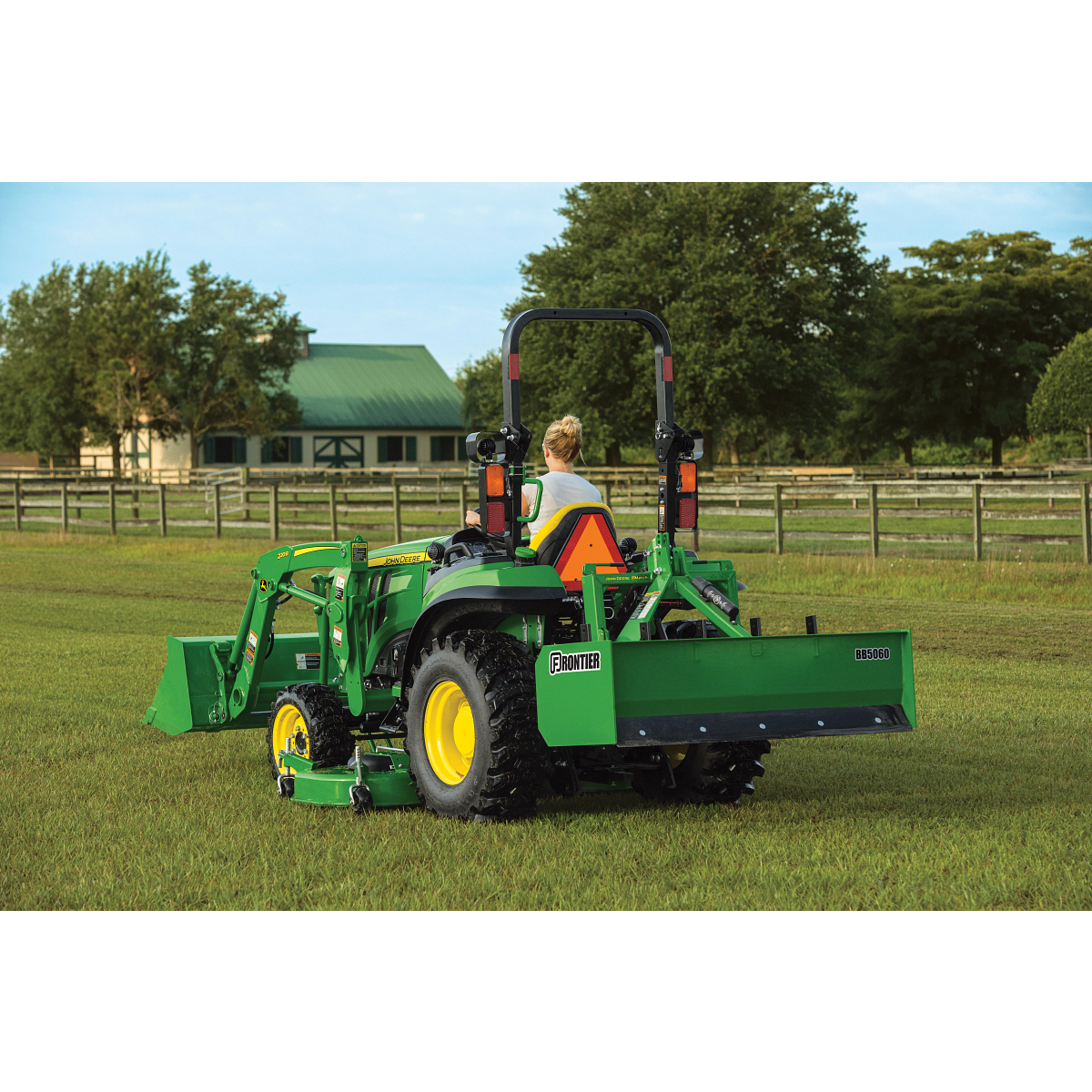 Green compact utility tractor with a front loader and rear implement attachment in a grassy field, surrounded by fencing and trees and farm buildings visible in the background