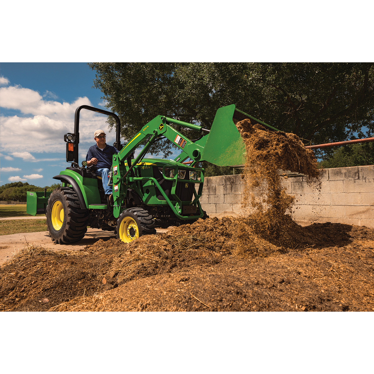 Green tractor with a front loader lifts and dumps a pile of fertilizer beside a concrete barrier, with loose soil scattered on the ground. Trees and a bright blue sky with clouds are visible in the background.