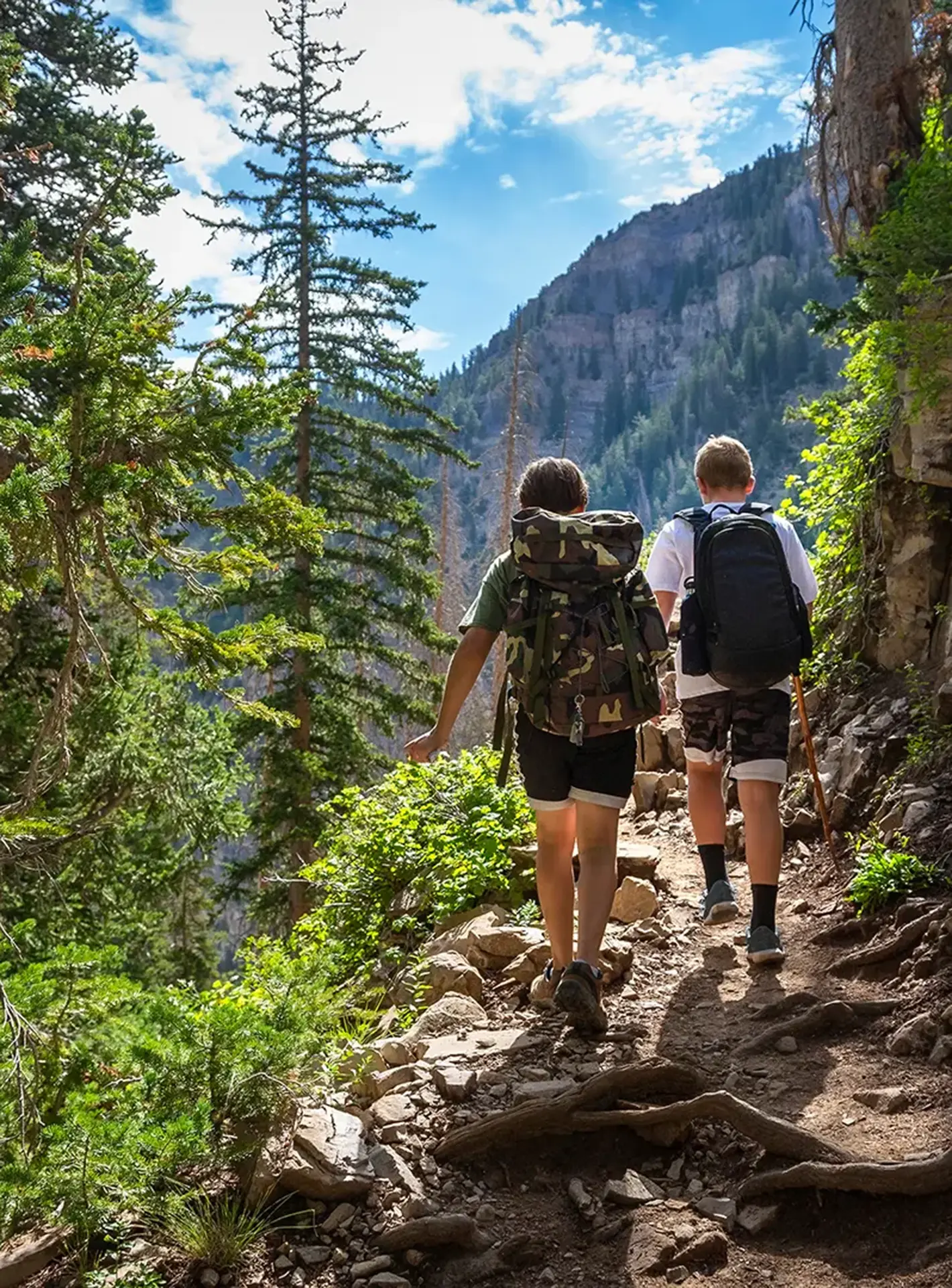 Two boys hiking on a rocky mountain trail through pine forest with backpacks, enjoying a sunny day in the Wasatch Mountains.